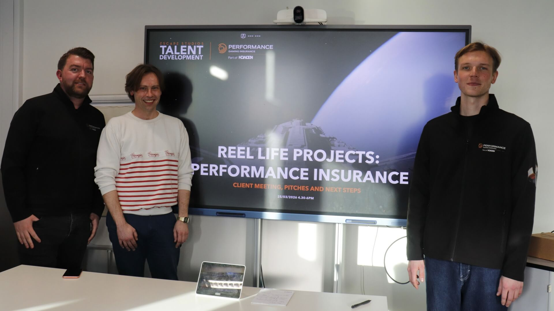 Project Leader Will Hughes from Escape Studios and Colin Powell and Gustaf Smith from Howden Performance standing next to a screen showing a presentation slide. The slide reads: Reel Life Projects: Performance Insurance.